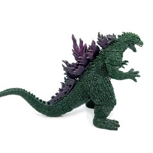K&ouml;nnte beinhalten: Eine gr&uuml;ne Godzilla-Figur mit lila R&uuml;ckenflossen. Die Figur befindet sich in einer dynamischen Pose, mit offenem Mund und gebogenem Schwanz. Das Spielzeug ist aus Kunststoff und steht auf wei&szlig;em Hintergrund.