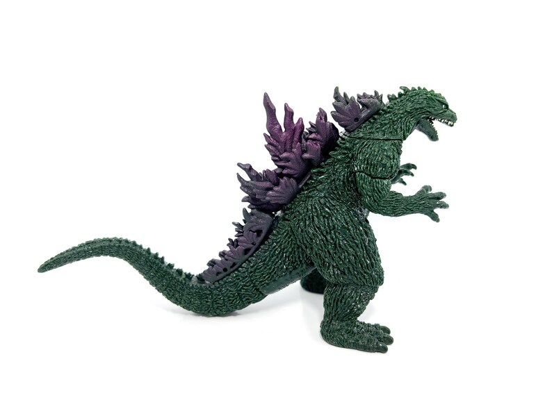 K&ouml;nnte beinhalten: Eine gr&uuml;ne Godzilla-Figur mit lila R&uuml;ckenflossen. Die Figur befindet sich in einer dynamischen Pose, mit offenem Mund und gebogenem Schwanz. Das Spielzeug ist aus Kunststoff und steht auf wei&szlig;em Hintergrund.