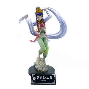May include: A figurine of a woman in a green and red outfit with a purple crown and a white scarf. She is standing on a cloud-shaped base with a black base that says "Lakshmi" in Japanese and English.