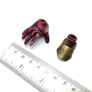 May include: Two action figure accessories: a red and gold hand and a gold forearm armor piece. The hand is open and the forearm armor is closed. A ruler is in the background for scale.