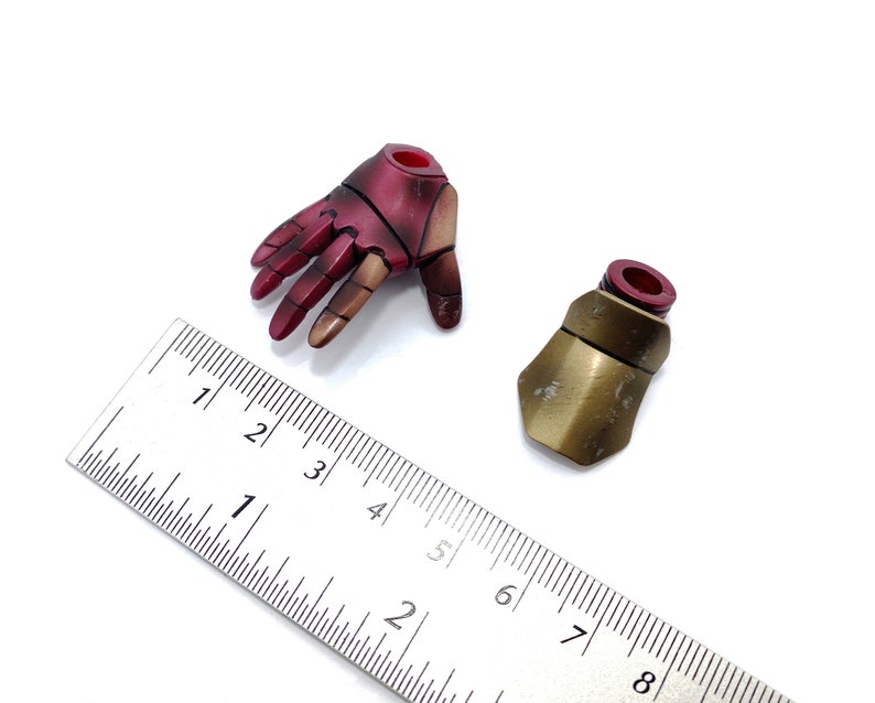 May include: Two action figure accessories: a red and gold hand and a gold forearm armor piece. The hand is open and the forearm armor is closed. A ruler is in the background for scale.