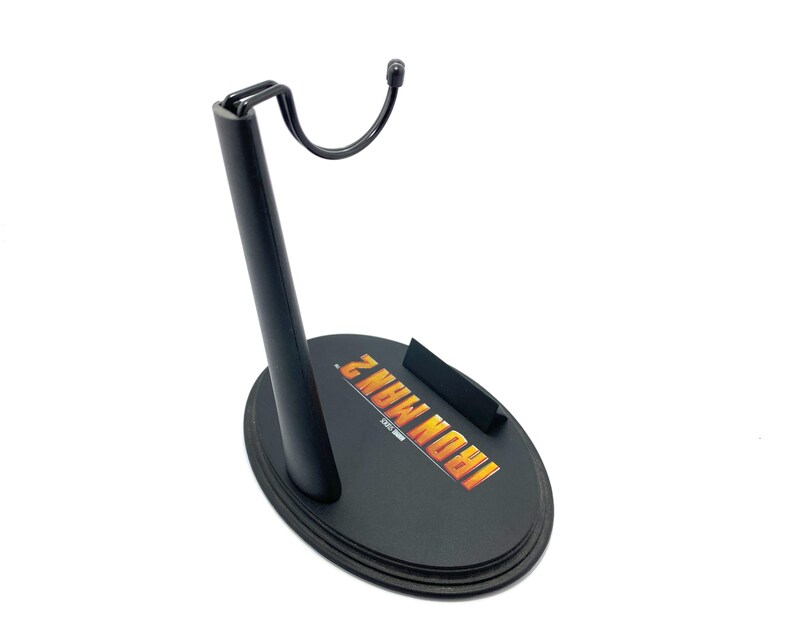 May include: Black plastic action figure stand with a hook and a circular base. The base has the words "IRON MAN 2" printed on it in yellow and orange.