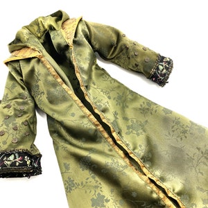 Op de afbeelding: Een lange, groene satijnen robe met een bloemenpatroon. De robe heeft gouden bies en decoratieve knopen. De mouwen hebben zwarte en gouden bies met kleurrijke borduurwerk.