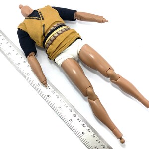 May include: A nude-toned articulated action figure with a black and gold shirt and white underwear. The figure is lying on its back with its arms outstretched and legs bent at the knees. A ruler is placed next to the figure for scale.