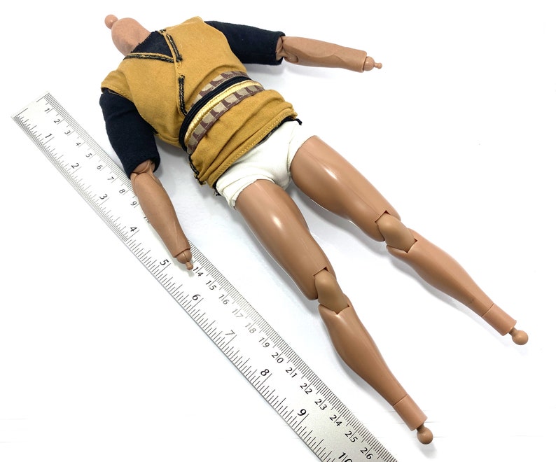 May include: A nude-toned articulated action figure with a black and gold shirt and white underwear. The figure is lying on its back with its arms outstretched and legs bent at the knees. A ruler is placed next to the figure for scale.