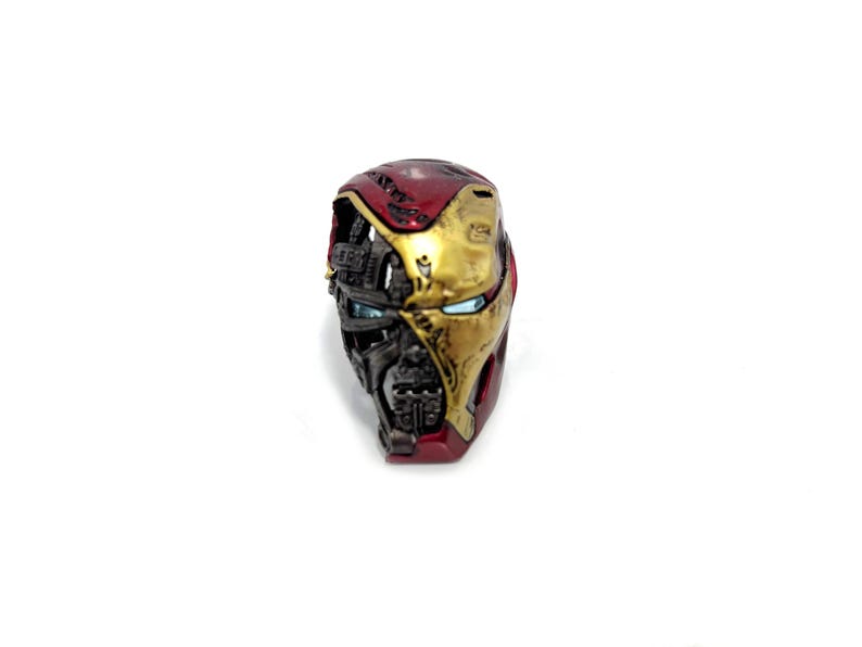 May include: A detailed, half-face replica of an Iron Man helmet. The helmet features a red and gold color scheme, with intricate mechanical detailing and visible internal components. The eye lenses are a bright blue.