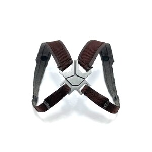 May include: A brown and gray harness with a silver-colored metal centerpiece. The harness has two shoulder straps and a connecting piece with a geometric design. The straps are made of a woven material.