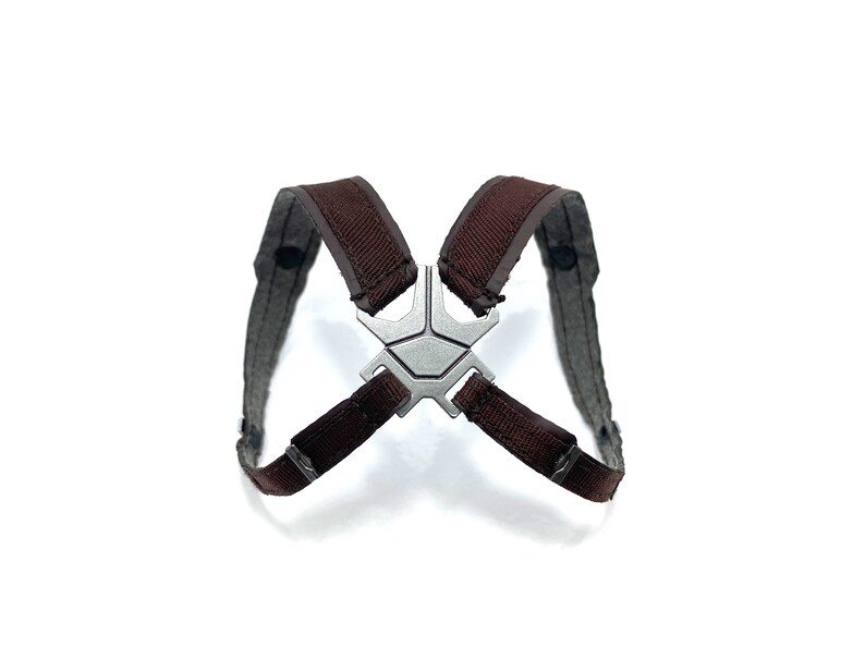 May include: A brown and gray harness with a silver-colored metal centerpiece. The harness has two shoulder straps and a connecting piece with a geometric design. The straps are made of a woven material.