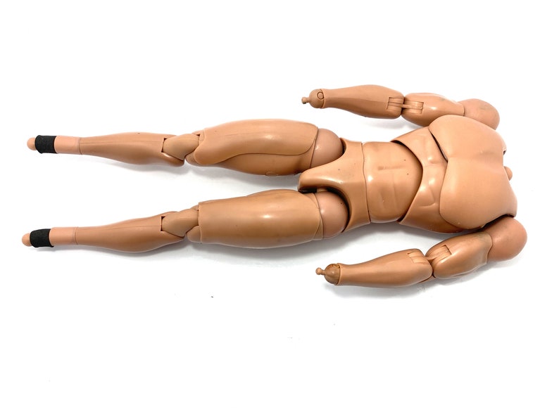 May include: A tan-colored articulated action figure body, lying on a white surface. The figure has jointed arms and legs, with black bands around the ankles. The figure is designed to be posed.