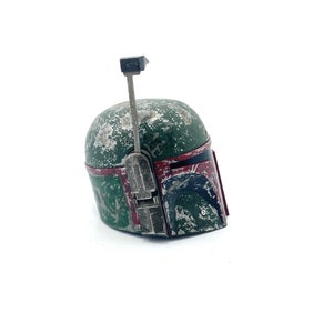May include: A weathered green and red Boba Fett helmet with a black antenna. The helmet has a worn and distressed look, with visible scratches and scuffs.