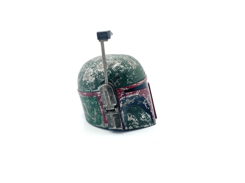 May include: A weathered green and red Boba Fett helmet with a black antenna. The helmet has a worn and distressed look, with visible scratches and scuffs.