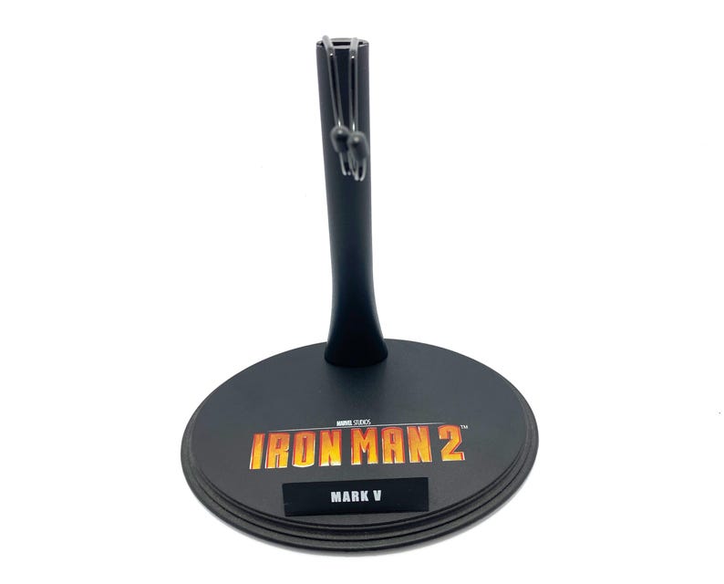 May include: Black plastic action figure stand with a round base. The base has the text "IRON MAN 2" in orange and yellow and "MARK V" in black.