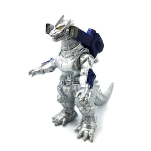 May include: A silver-colored robot toy with a reptilian head and tail, featuring blue and black accents. The toy has a mechanical design with visible joints and a backpack-like structure. The robot is standing on a white surface.