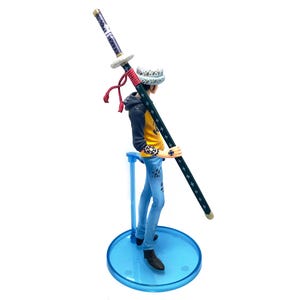 Pu&ograve; includere: Statuetta dettagliata di un personaggio che tiene una lunga spada. La figura indossa una giacca gialla e nera, jeans blu e un cappello fantasia. La spada &egrave; nera con stelle bianche e un'impugnatura viola. La statuetta &egrave; su un supporto blu.