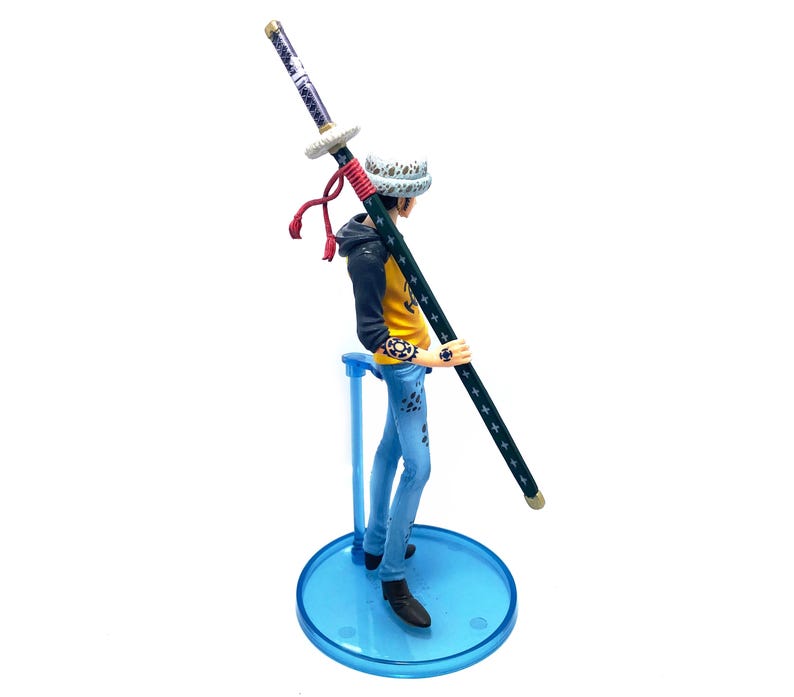 Pu&ograve; includere: Statuetta dettagliata di un personaggio che tiene una lunga spada. La figura indossa una giacca gialla e nera, jeans blu e un cappello fantasia. La spada &egrave; nera con stelle bianche e un'impugnatura viola. La statuetta &egrave; su un supporto blu.