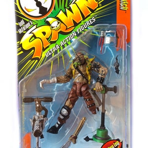 May include: A green, yellow, and black Spawn action figure with a green, yellow, and black packaging. The figure is holding a green and black weapon. The packaging has the words "Todd McFarlane's Spawn Ultra-Action Figures" and "Fires Peg Leg Machele!" on it.