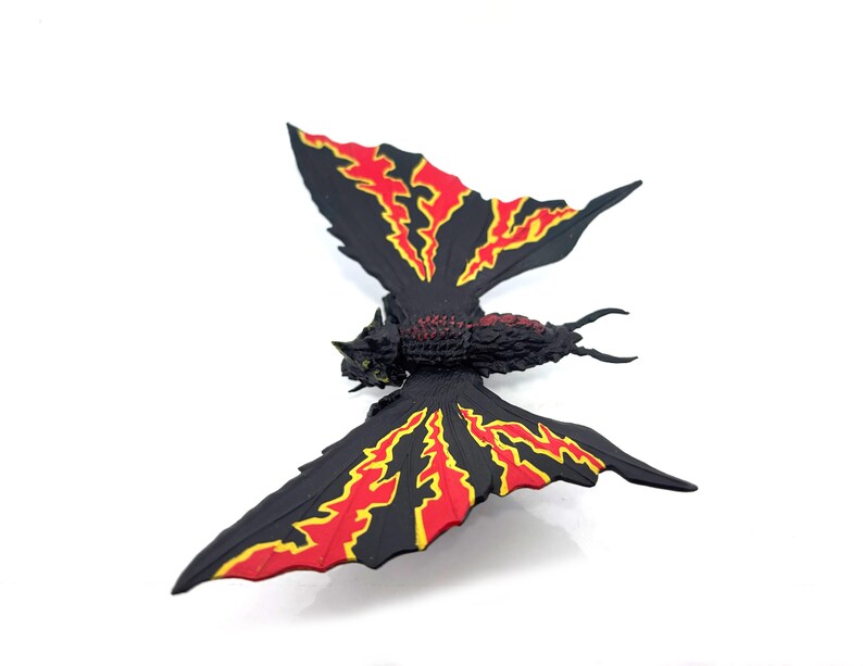 May include: A black insect-like figurine with a textured body and two large wings. The wings feature a jagged, flame-like pattern in bright red and yellow against a solid black background. The creature has a segmented torso and two thin antennae.