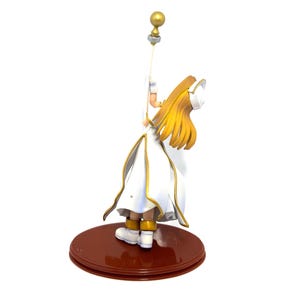 May include: A figurine of a character in a white and gold outfit, holding a staff with a golden orb. The figure has long, flowing blonde hair and is standing on a brown, circular base. The outfit has gold trim.