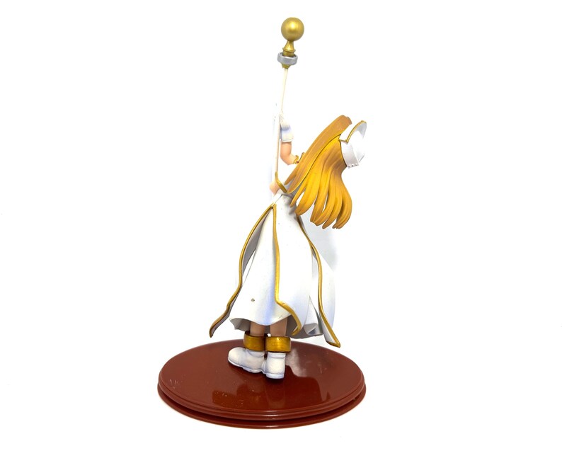 May include: A figurine of a character in a white and gold outfit, holding a staff with a golden orb. The figure has long, flowing blonde hair and is standing on a brown, circular base. The outfit has gold trim.