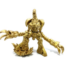 May include: A gold-coloured, articulated action figure of a creature with a humanoid torso, large clawed hands, and a spiky base. The figure has a detailed, textured surface and is posed on a white background.