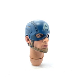 Op de afbeelding: Een gedetailleerde hoofdsnede van Captain America, met een blauwe helm met een wit "A"-embleem. Het gezicht heeft realistische huidtinten en blauwe ogen. Het hoofd is bevestigd aan een bruine nek en schouders, met een bruine kinriem.