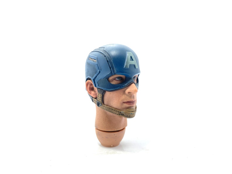 Op de afbeelding: Een gedetailleerde hoofdsnede van Captain America, met een blauwe helm met een wit "A"-embleem. Het gezicht heeft realistische huidtinten en blauwe ogen. Het hoofd is bevestigd aan een bruine nek en schouders, met een bruine kinriem.