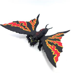 May include: A black insectoid figurine with large, spread wings featuring jagged red and yellow patterns. The creature has a textured, dark body with a small head, multiple legs, and two antennae, captured from a high-angle perspective.
