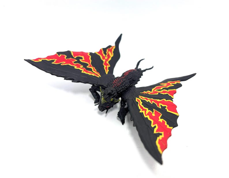 May include: A black insectoid figurine with large, spread wings featuring jagged red and yellow patterns. The creature has a textured, dark body with a small head, multiple legs, and two antennae, captured from a high-angle perspective.