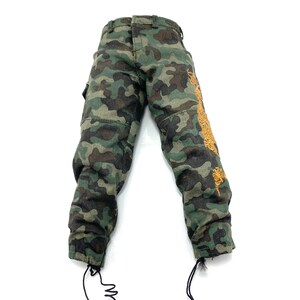 May include: Camouflage trousers with a brown and green pattern. The trousers have a drawstring at the bottom of each leg and a gold embroidered design on the right leg.