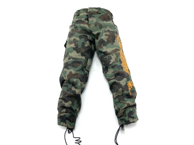 May include: Camouflage trousers with a brown and green pattern. The trousers have a drawstring at the bottom of each leg and a gold embroidered design on the right leg.