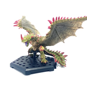 Op de afbeelding: Gedetailleerd beeldje van een draak met groene, bruine en rode accenten. De draak heeft grote vleugels, een stekelige staart en is gemonteerd op een zwarte zeshoekige basis. De bek van de draak is open en onthult paarse tanden.