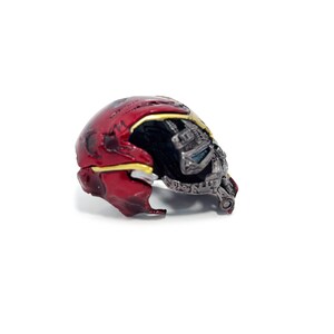 May include: A detailed, miniature replica of a red and gold helmet, with a partially exposed metallic faceplate. The helmet features intricate detailing, including a yellow trim and a dark interior, set against a white background.