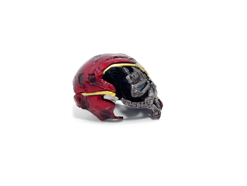 May include: A detailed, miniature replica of a red and gold helmet, with a partially exposed metallic faceplate. The helmet features intricate detailing, including a yellow trim and a dark interior, set against a white background.