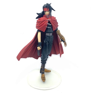 May include: A black and red action figure of a character wearing a long red cape and holding a gun. The figure is standing on a clear plastic base.