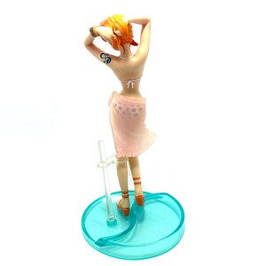May include: A figurine of a woman with orange hair wearing a pink sarong and a white bikini top. She is standing on a blue and clear plastic base.