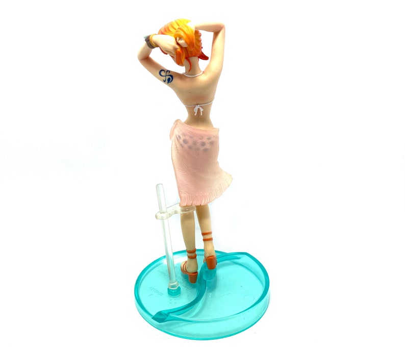 May include: A figurine of a woman with orange hair wearing a pink sarong and a white bikini top. She is standing on a blue and clear plastic base.
