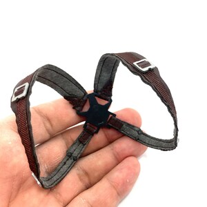 May include: A brown and gray harness with silver-colored buckles and a black central connector. The harness is made of a woven material and is designed to be worn on the body.