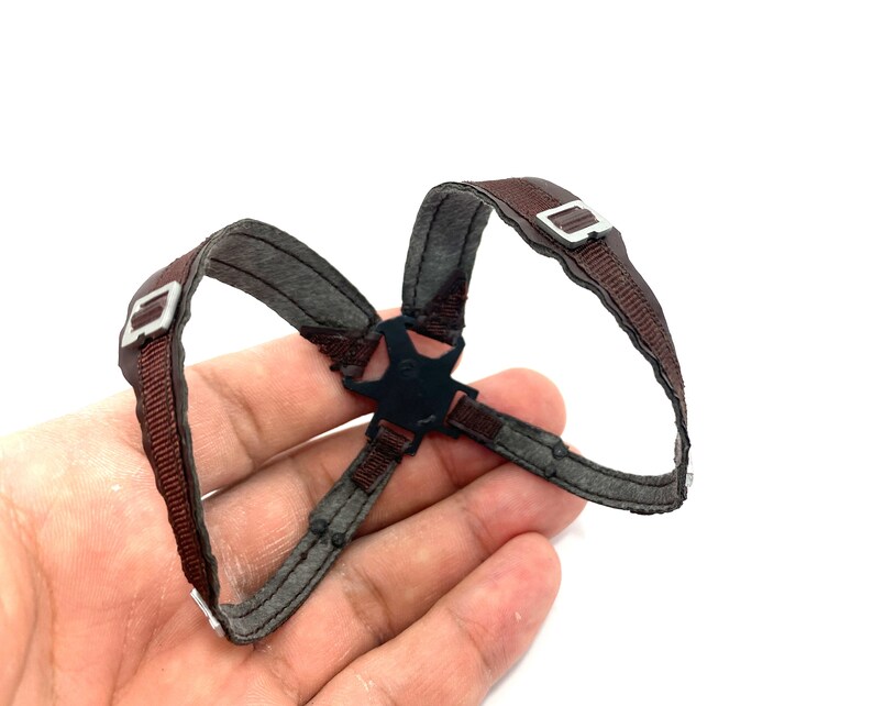 May include: A brown and gray harness with silver-colored buckles and a black central connector. The harness is made of a woven material and is designed to be worn on the body.