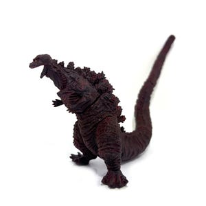 May include: A maroon Godzilla figurine with a textured, spiky back and a long tail. The figure is posed with its mouth open, showcasing its teeth. The toy is set against a white background.
