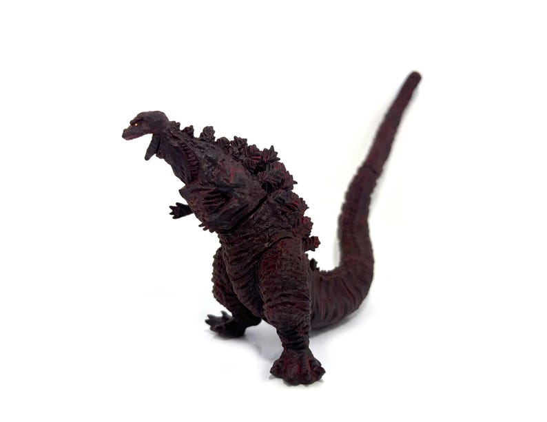 May include: A maroon Godzilla figurine with a textured, spiky back and a long tail. The figure is posed with its mouth open, showcasing its teeth. The toy is set against a white background.