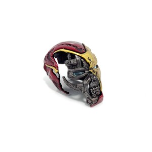 May include: A detailed, partially disassembled Iron Man helmet, showcasing its internal mechanics. The helmet features a red and gold color scheme, with visible mechanical components and intricate detailing. The design is set against a plain white background.