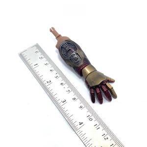K&ouml;nnte beinhalten: Detaillierter Actionfigurenarm mit metallischer Gold- und Burgunderfarbgebung. Der Arm ist etwa 13 cm lang und weist komplizierte mechanische Details auf. Ein Lineal liegt zur Ma&szlig;stabsbestimmung neben dem Arm.