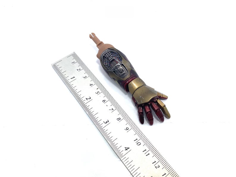 K&ouml;nnte beinhalten: Detaillierter Actionfigurenarm mit metallischer Gold- und Burgunderfarbgebung. Der Arm ist etwa 13 cm lang und weist komplizierte mechanische Details auf. Ein Lineal liegt zur Ma&szlig;stabsbestimmung neben dem Arm.