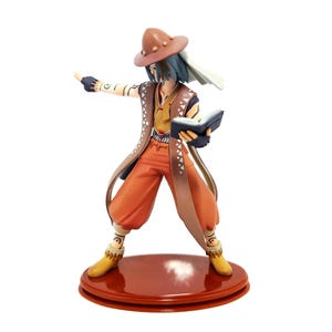 May include: A figurine of a character in a brown hat, orange pants, and a long brown coat. The character is holding a book and pointing with one hand. The figurine is standing on a round, reddish-brown base.