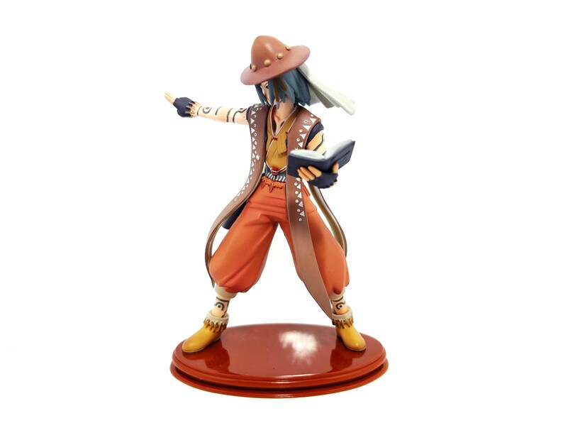May include: A figurine of a character in a brown hat, orange pants, and a long brown coat. The character is holding a book and pointing with one hand. The figurine is standing on a round, reddish-brown base.