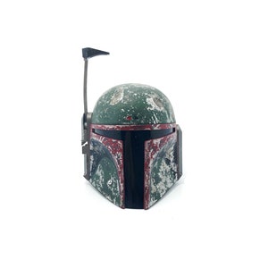 May include: A green and red plastic Boba Fett helmet with a weathered and worn look. The helmet has a black visor and a small antenna on top.