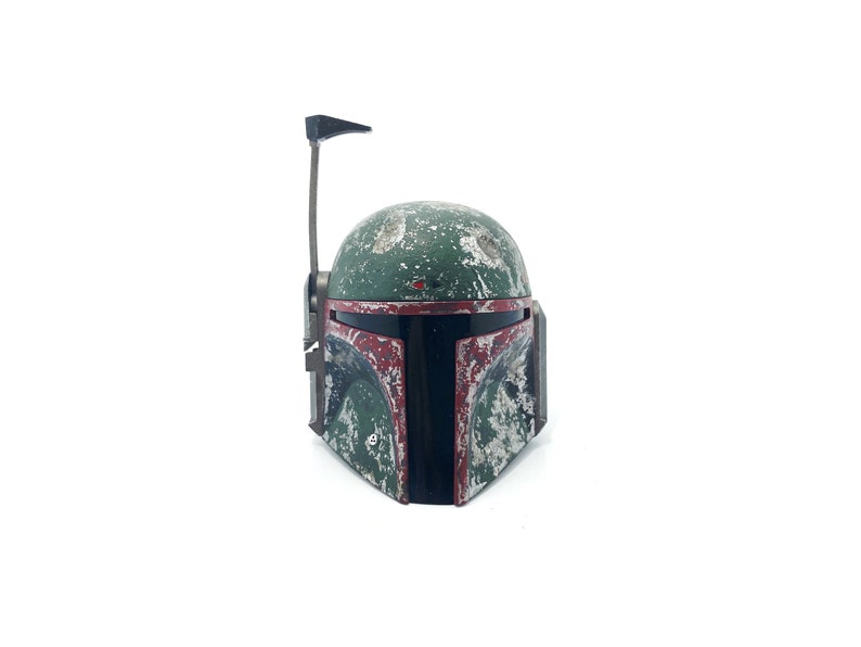May include: A green and red plastic Boba Fett helmet with a weathered and worn look. The helmet has a black visor and a small antenna on top.