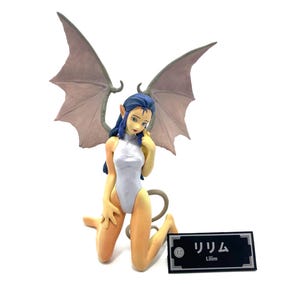 Puede incluir: Una figura de una demonio femenina con cabello azul y alas grises. Lleva un traje de baño blanco de una pieza y está arrodillada con la cabeza inclinada hacia abajo. La figura está etiquetada como "Lilim" en japonés e inglés.