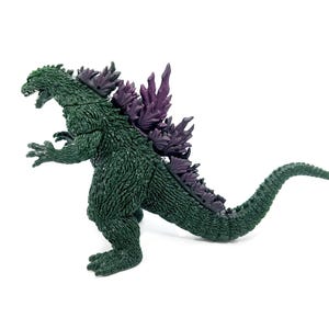 K&ouml;nnte beinhalten: Eine gr&uuml;ne Godzilla-Actionfigur mit lila R&uuml;ckenflossen und Stacheln. Der Spielzeug-Dinosaurier ist in einer dynamischen Haltung dargestellt, mit offenem Maul und gebogenem Schwanz. Die Figur befindet sich auf wei&szlig;em Hintergrund.