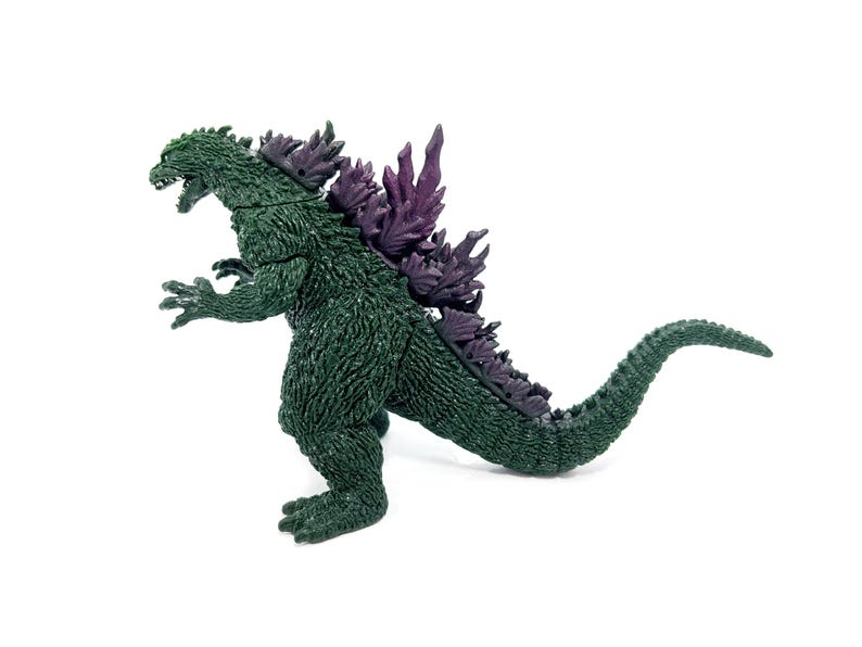 K&ouml;nnte beinhalten: Eine gr&uuml;ne Godzilla-Actionfigur mit lila R&uuml;ckenflossen und Stacheln. Der Spielzeug-Dinosaurier ist in einer dynamischen Haltung dargestellt, mit offenem Maul und gebogenem Schwanz. Die Figur befindet sich auf wei&szlig;em Hintergrund.